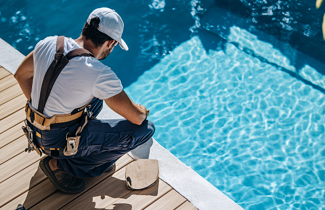 A worker in overalls and a white cap kneels by the edge of a swimming pool, examining the poolside deck in daylight.