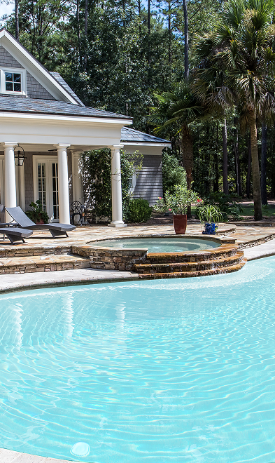 A backyard with a swimming pool, raised hot tub, lounge chairs, plants, and a house with columns surrounded by trees.