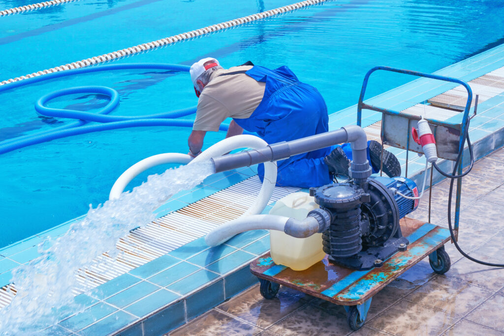 A worker in blue overalls operates pool cleaning equipment beside a swimming pool, with water being pumped out through a hose.