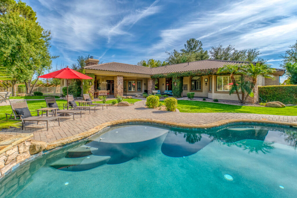 A backyard features a pool, patio furniture with a red umbrella, green lawn, and a single-story house with a covered porch.