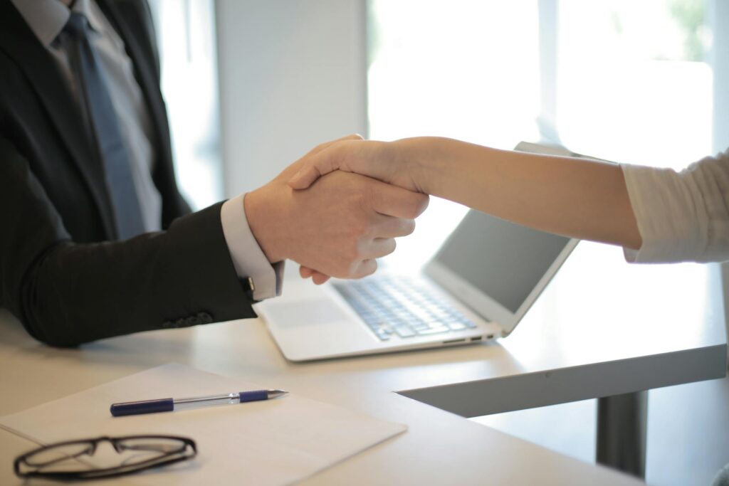 Two people shake hands across a desk with a laptop, pen, and paper visible in an office setting.