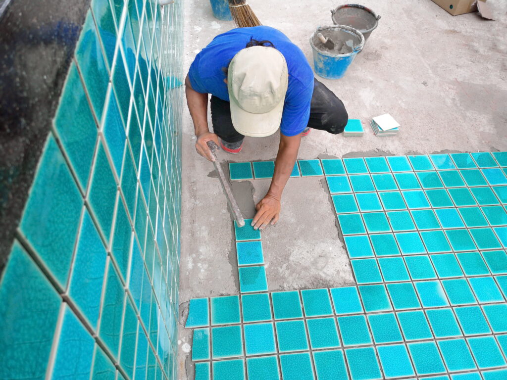A person wearing a blue shirt and beige cap installs blue square tiles on a floor with trowel and adhesive.
