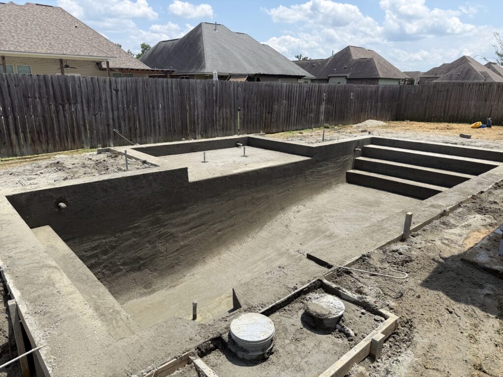 Rectangular in-ground swimming pool under construction with concrete shell, steps on one end, and a fenced backyard.