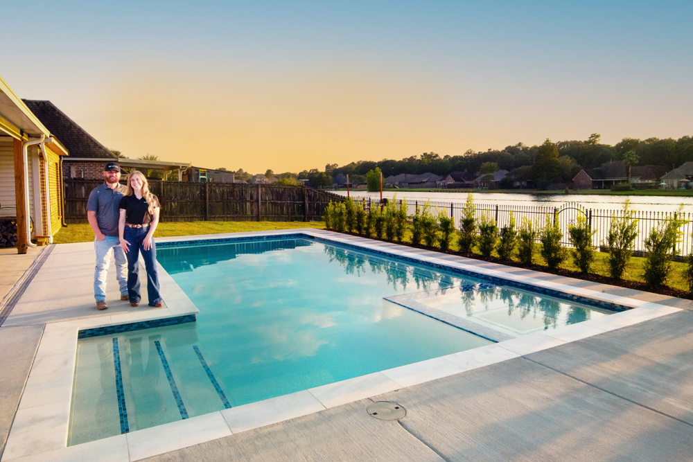 A man and woman stand by a rectangular backyard pool with landscaping, a fence, and a pond and houses in the background at sunset.