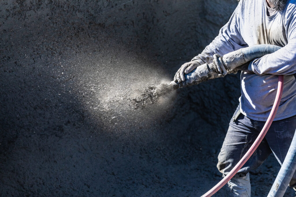Person wearing protective clothing spraying concrete onto a surface with a hose, creating a fine mist of concrete particles.