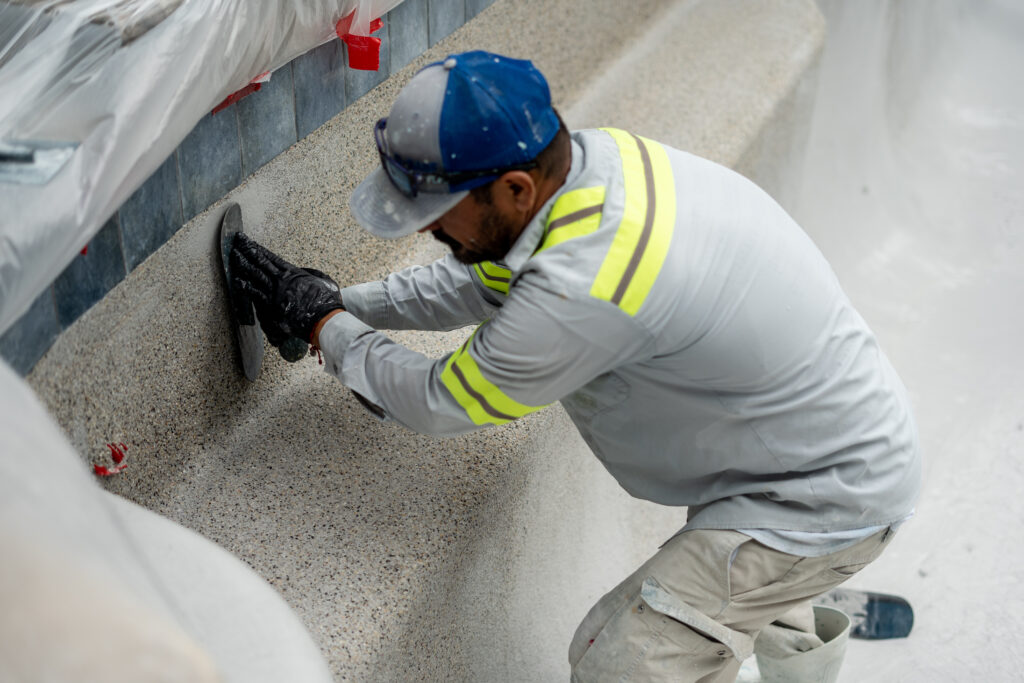 A worker in safety gear smooths the edge of a concrete surface with a hand tool inside an unfinished pool.