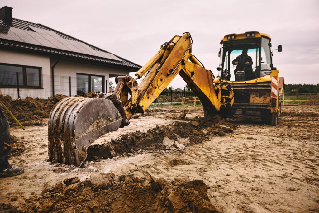 A yellow backhoe loader digging and moving earth at a construction site near a modern house.