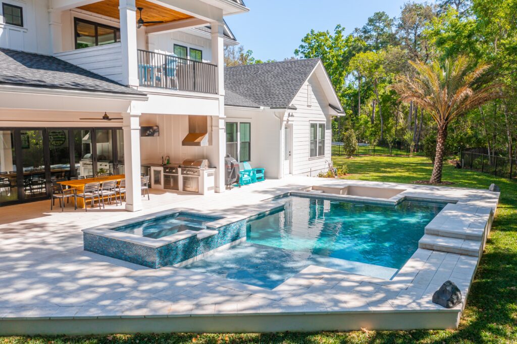 Modern backyard with a rectangular pool, hot tub, outdoor kitchen, dining area, and surrounding trees on a sunny day.