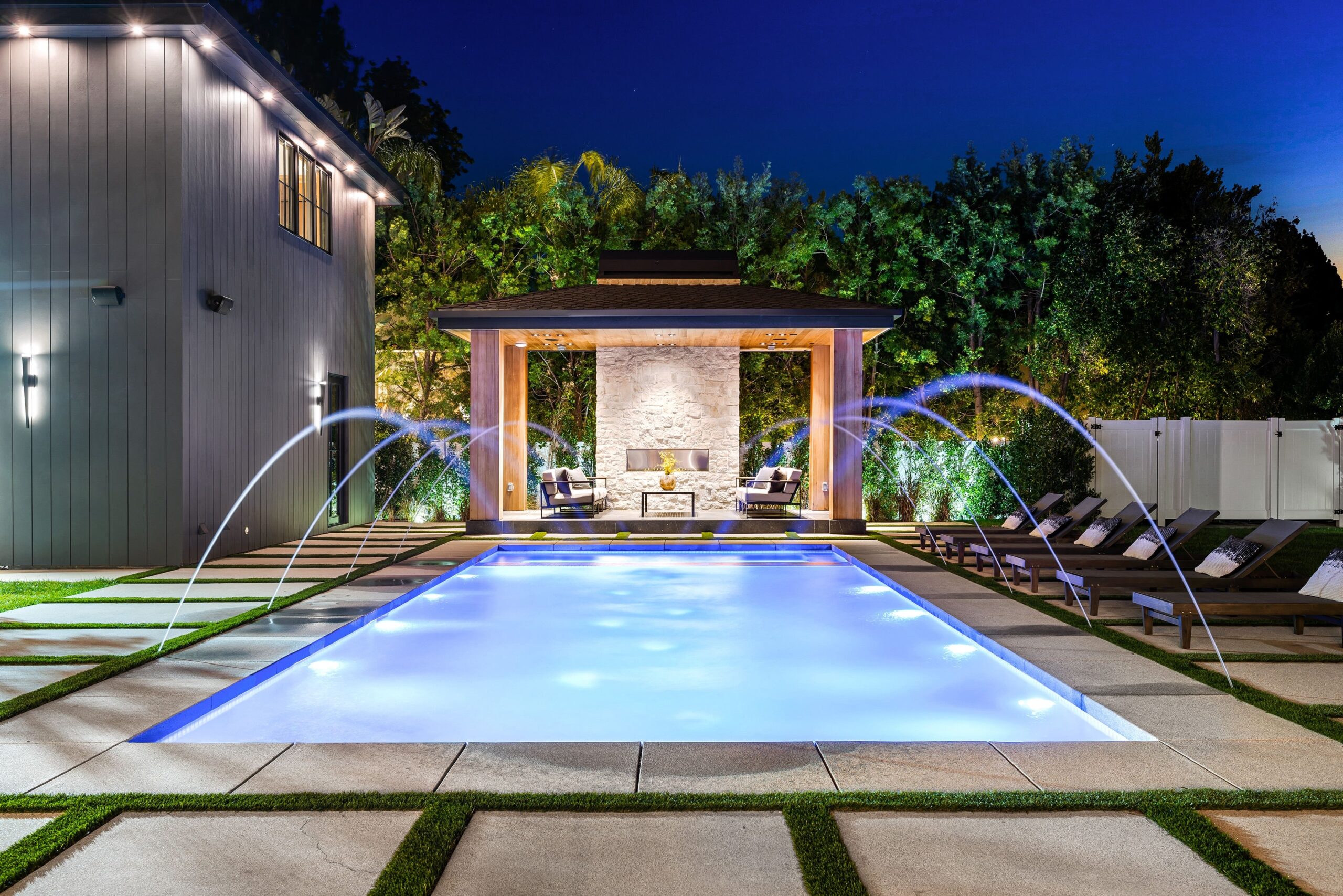 A modern backyard pool at night with water jets, lounge chairs, and a covered seating area with a fireplace in the background.